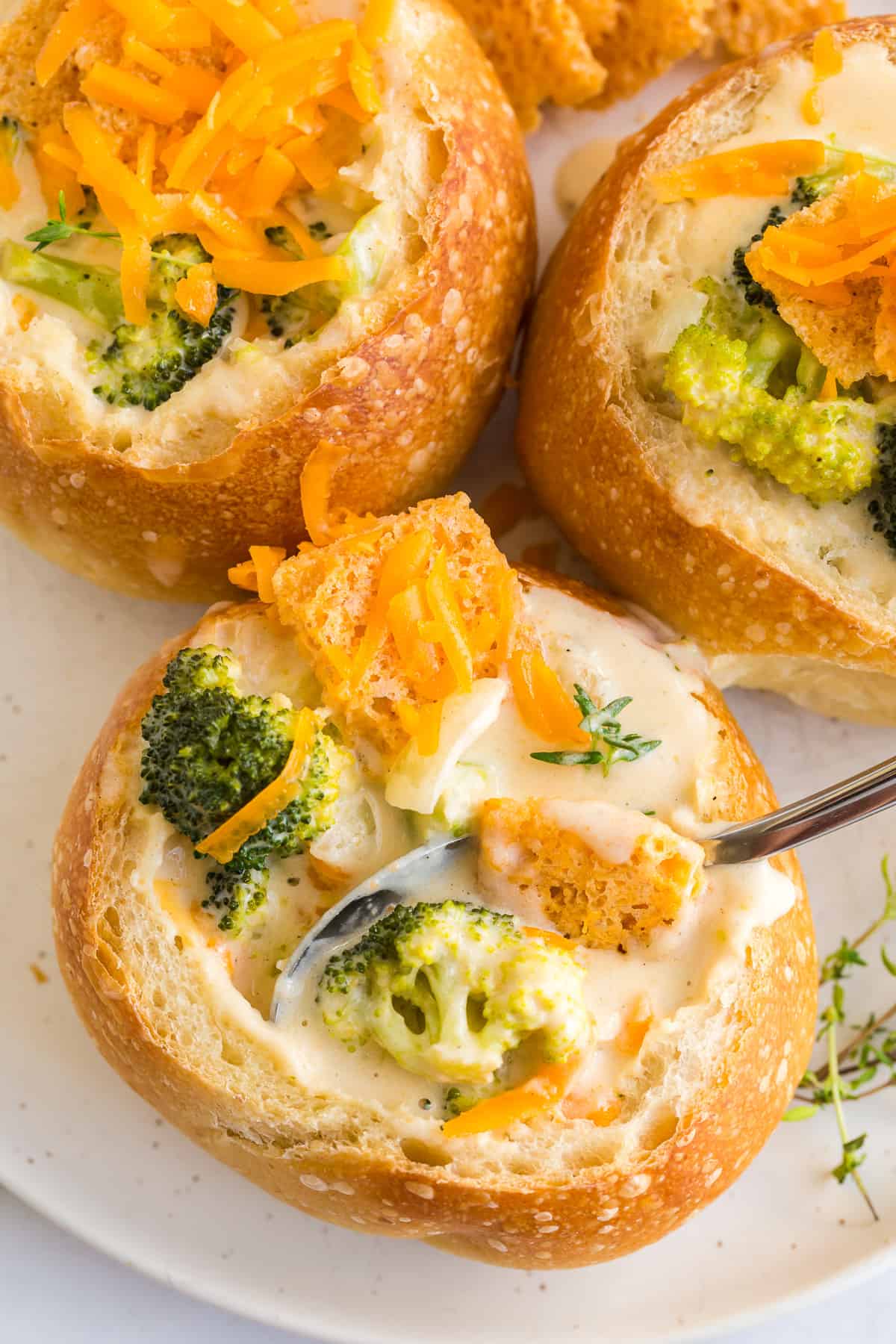 close up overhead view of broccoli cheddar soup topped with fresh thyme, grated cheddar cheese, and a cheese cracker served in a sourdough bread bowl