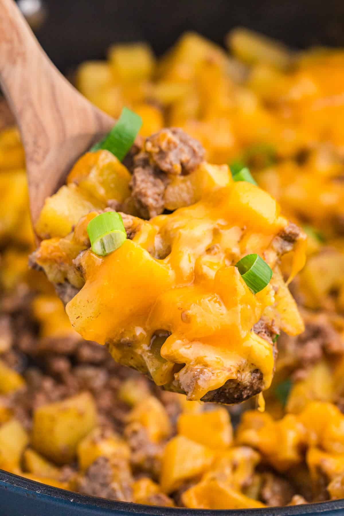 Close-up of Cheesy Ground Beef and Potato Skillet on a wooden spoon with diced Yukon Gold potatoes, ground beef, melted cheddar cheese, and green onions.