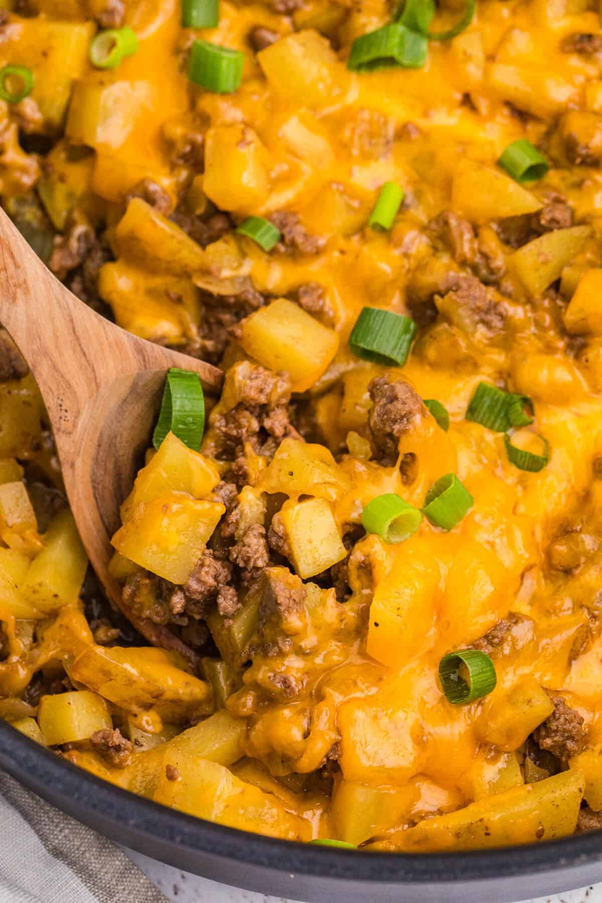 Close-up of Cheesy Ground Beef and Potato Skillet with diced Yukon Gold potatoes, seasoned ground beef, melted cheddar cheese, and green onions being scooped up with a wooden spoon.