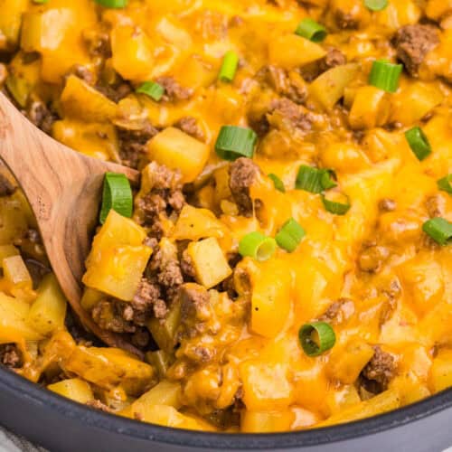 Close-up of Cheesy Ground Beef and Potato Skillet with diced Yukon Gold potatoes, seasoned ground beef, melted cheddar cheese, and green onions in a black skillet with a wooden spoon.